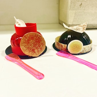 Two intricately designed desserts sit on a white surface. The dessert on the left is vibrant red with a round decorative disk on the side and a small white cream accent on top. The dessert on the right features a dark, glossy dome with a textured white and brown circular accent, topped with a white and brown speckled piece. Each dessert is served on a black base with a pink plastic spoon placed alongside.