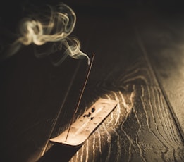 A lit incense stick emits a gentle wisp of smoke, placed on a wooden holder. The background is a dark wooden surface, which adds a warm and cozy atmosphere.