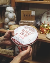 A person is holding a round package of Chinese tea with Chinese characters written on it. The background is filled with shelves holding various tea products and a gold-colored decorative item.