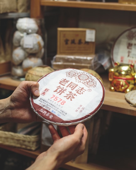 A person is holding a round package of Chinese tea with Chinese characters written on it. The background is filled with shelves holding various tea products and a gold-colored decorative item.