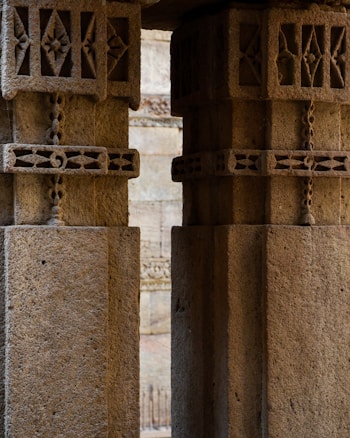 Intricate stone columns with decorative carvings and geometric patterns, set in a historical architectural structure. The texture of the stone is rough with visible details, suggesting age and craftsmanship.
