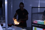 A person is cooking at a food stall, using a high flame to prepare a dish. The setting seems to be indoors, possibly in a market or fair. Various kitchen items and ingredients are visible around, including bowls and packaged products.