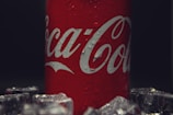 A red can with white lettering featuring water droplets, surrounded by ice cubes. The background is dark, highlighting the can.