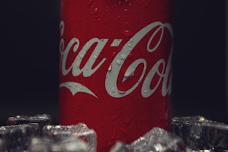 A red can with white lettering featuring water droplets, surrounded by ice cubes. The background is dark, highlighting the can.