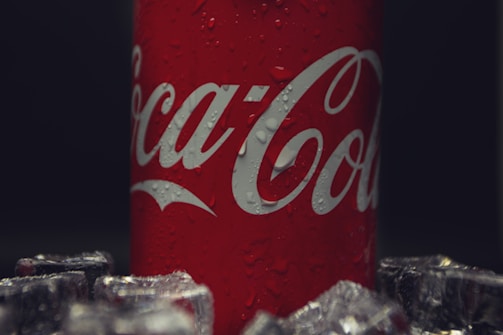 A red can with white lettering featuring water droplets, surrounded by ice cubes. The background is dark, highlighting the can.