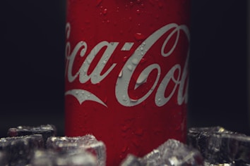 A red can with white lettering featuring water droplets, surrounded by ice cubes. The background is dark, highlighting the can.