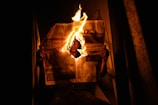 A person sitting in a dark area holding a newspaper that is partially on fire, with flames prominently rising in the center. The light from the fire illuminates parts of the scene, creating a stark contrast against the surrounding darkness.