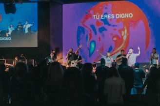 A group of musicians and singers perform on a stage with an audience in front, energized and engaged. The stage is illuminated with a vibrant, abstract visual projection that displays the phrase 'TU ERES DIGNO.' The lighting creates a lively atmosphere with a focus on red and blue tones.