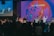 A group of musicians and singers perform on a stage with an audience in front, energized and engaged. The stage is illuminated with a vibrant, abstract visual projection that displays the phrase 'TU ERES DIGNO.' The lighting creates a lively atmosphere with a focus on red and blue tones.