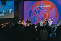A group of musicians and singers perform on a stage with an audience in front, energized and engaged. The stage is illuminated with a vibrant, abstract visual projection that displays the phrase 'TU ERES DIGNO.' The lighting creates a lively atmosphere with a focus on red and blue tones.