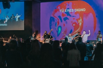 A group of musicians and singers perform on a stage with an audience in front, energized and engaged. The stage is illuminated with a vibrant, abstract visual projection that displays the phrase 'TU ERES DIGNO.' The lighting creates a lively atmosphere with a focus on red and blue tones.