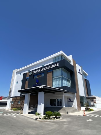 A modern building with sleek architecture featuring large glass windows and a blend of white and dark exterior sections. The structure is labeled 'Center of Excellence'. There are stairs and a ramp at the entrance, and the surrounding area is paved, with some minimal landscaping including small plants.