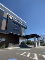 A modern building with a sleek, contemporary design prominently features the words 'Center of Excellence' on its facade. The structure has large glass windows and dark panels, and a covered entrance. The surrounding area includes a parking lot with visible handicap spaces and some greenery, such as small bushes and trees.