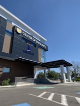 A modern building with a sleek, contemporary design prominently features the words 'Center of Excellence' on its facade. The structure has large glass windows and dark panels, and a covered entrance. The surrounding area includes a parking lot with visible handicap spaces and some greenery, such as small bushes and trees.