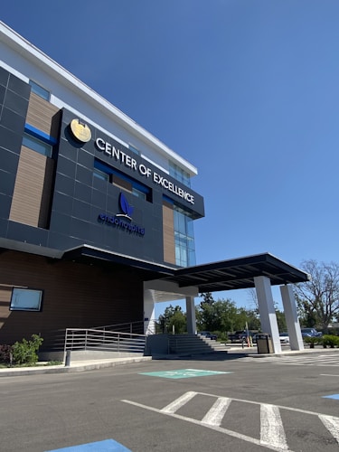 A modern building with a sleek, contemporary design prominently features the words 'Center of Excellence' on its facade. The structure has large glass windows and dark panels, and a covered entrance. The surrounding area includes a parking lot with visible handicap spaces and some greenery, such as small bushes and trees.