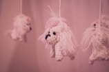 Hanging crochet toys resembling small white dogs with long fur and pink bows on their heads, suspended from strings against a soft pink background.