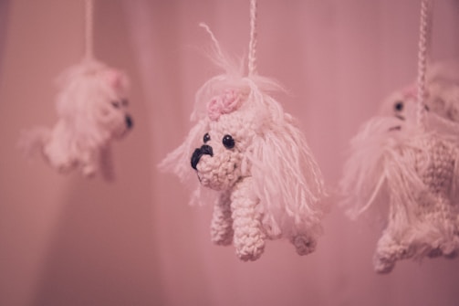 Hanging crochet toys resembling small white dogs with long fur and pink bows on their heads, suspended from strings against a soft pink background.