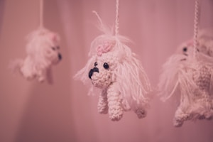 Hanging crochet toys resembling small white dogs with long fur and pink bows on their heads, suspended from strings against a soft pink background.