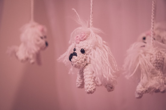 Hanging crochet toys resembling small white dogs with long fur and pink bows on their heads, suspended from strings against a soft pink background.