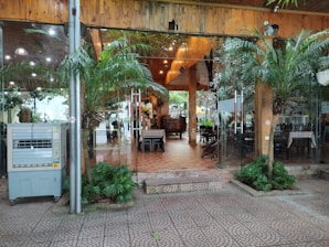 Glass doors with metal handles leading to an indoor area with wooden ceilings and tiled floors. There are tables and chairs visible inside, surrounded by decorative plants both outside and inside. An air conditioning unit is positioned beside a metallic pole near the entrance.