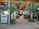 Glass doors with metal handles leading to an indoor area with wooden ceilings and tiled floors. There are tables and chairs visible inside, surrounded by decorative plants both outside and inside. An air conditioning unit is positioned beside a metallic pole near the entrance.