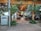 Glass doors with metal handles leading to an indoor area with wooden ceilings and tiled floors. There are tables and chairs visible inside, surrounded by decorative plants both outside and inside. An air conditioning unit is positioned beside a metallic pole near the entrance.