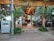 Glass doors with metal handles leading to an indoor area with wooden ceilings and tiled floors. There are tables and chairs visible inside, surrounded by decorative plants both outside and inside. An air conditioning unit is positioned beside a metallic pole near the entrance.