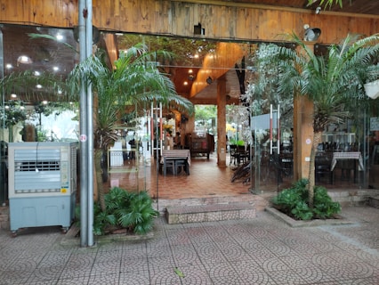 Glass doors with metal handles leading to an indoor area with wooden ceilings and tiled floors. There are tables and chairs visible inside, surrounded by decorative plants both outside and inside. An air conditioning unit is positioned beside a metallic pole near the entrance.