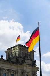 Two German flags flutter against a clear blue sky with scattered clouds. Below, a historic stone building features intricate architectural details and multiple windows.