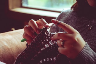 A person is engaged in a knitting activity, holding knitting needles and working with yarn. The close-up view captures the details of their hands and the fabric in progress. The setting appears cozy with natural light coming from a nearby window.