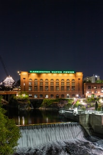 A well-lit, historic brick building with green neon lettering reads 'Washington Water Power.' The structure is situated along a river with a cascading waterfall. The night sky and city lights are visible in the background.