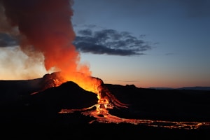 A volcanic eruption with bright, glowing lava flowing down the side of a mountain. The orange and red hues of the lava contrast against the dark silhouette of the surrounding landscape. Above, a plume of smoke rises into the sky, blending with the twilight atmosphere of deep blues and purples.