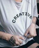 A person wearing a white T-shirt with the word 'CREATIVE' printed on it in black letters. The person's hands, featuring tattoos and a bracelet, are gripping the shirt.