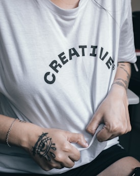 A person wearing a white T-shirt with the word 'CREATIVE' printed on it in black letters. The person's hands, featuring tattoos and a bracelet, are gripping the shirt.