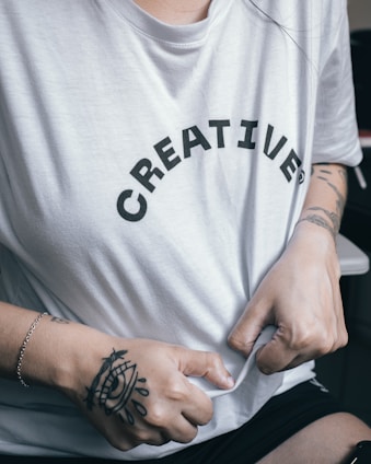 A person wearing a white T-shirt with the word 'CREATIVE' printed on it in black letters. The person's hands, featuring tattoos and a bracelet, are gripping the shirt.