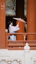 A person dressed in traditional Asian attire stands on a wooden terrace holding an ornate parasol. The scene is set against a backdrop of large red columns, with intricate decorative details on the railings. The person appears to be looking outwards, creating a serene and contemplative atmosphere.