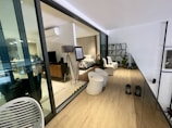 A modern, indoor living space featuring a bedroom visible through large glass sliding doors. The bedroom is furnished with a bed, bedside table, floor lamp, and a TV on a wooden stand. There is an easel with a framed piece of information. The adjacent area includes white chairs, round tables, and plants on a decorative wall grid. Two lanterns are placed on the wooden floor, and another living area is visible through the glass with more furniture and decor.