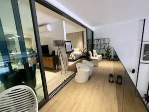A modern, indoor living space featuring a bedroom visible through large glass sliding doors. The bedroom is furnished with a bed, bedside table, floor lamp, and a TV on a wooden stand. There is an easel with a framed piece of information. The adjacent area includes white chairs, round tables, and plants on a decorative wall grid. Two lanterns are placed on the wooden floor, and another living area is visible through the glass with more furniture and decor.
