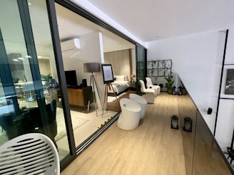 A modern, indoor living space featuring a bedroom visible through large glass sliding doors. The bedroom is furnished with a bed, bedside table, floor lamp, and a TV on a wooden stand. There is an easel with a framed piece of information. The adjacent area includes white chairs, round tables, and plants on a decorative wall grid. Two lanterns are placed on the wooden floor, and another living area is visible through the glass with more furniture and decor.
