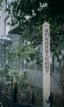 A vertical white post with black text stands surrounded by green foliage, next to a building with large windows. The ground has dried leaves and a small black fence.