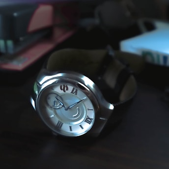 A stylish wristwatch with a silver casing and intricate Roman numerals on its dial, reflecting light in a dimly lit environment. The close-up perspective highlights its elegant design, with hints of surrounding objects blurred in the background.