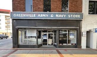 A storefront with a sign reading 'Greenville Army & Navy Store'. The facade features a brick and white exterior with a vintage appearance. The store's windows are partially covered with signs advertising for lease and tactical apparel. The street in front is paved, with a truck and some residential buildings visible on the left side.
