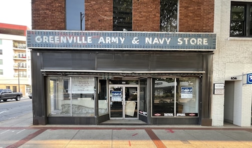 A storefront with a sign reading 'Greenville Army & Navy Store'. The facade features a brick and white exterior with a vintage appearance. The store's windows are partially covered with signs advertising for lease and tactical apparel. The street in front is paved, with a truck and some residential buildings visible on the left side.