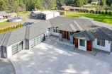 A modern house with a large driveway, featuring a sleek design with a mix of white and dark exterior finishes. The roof is multi-tiered, and large windows are visible. There are landscaped areas around the property, including a lawn and various outbuildings in the background.
