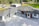 A modern house with a large driveway, featuring a sleek design with a mix of white and dark exterior finishes. The roof is multi-tiered, and large windows are visible. There are landscaped areas around the property, including a lawn and various outbuildings in the background.