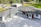 A modern house with a large driveway, featuring a sleek design with a mix of white and dark exterior finishes. The roof is multi-tiered, and large windows are visible. There are landscaped areas around the property, including a lawn and various outbuildings in the background.