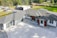 A modern house with a large driveway, featuring a sleek design with a mix of white and dark exterior finishes. The roof is multi-tiered, and large windows are visible. There are landscaped areas around the property, including a lawn and various outbuildings in the background.