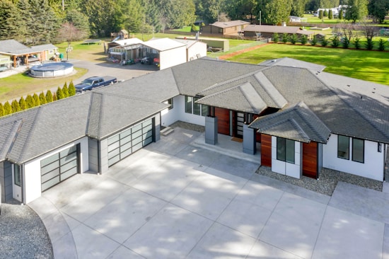 A modern house with a large driveway, featuring a sleek design with a mix of white and dark exterior finishes. The roof is multi-tiered, and large windows are visible. There are landscaped areas around the property, including a lawn and various outbuildings in the background.