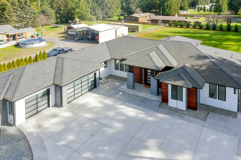 A modern house with a large driveway, featuring a sleek design with a mix of white and dark exterior finishes. The roof is multi-tiered, and large windows are visible. There are landscaped areas around the property, including a lawn and various outbuildings in the background.