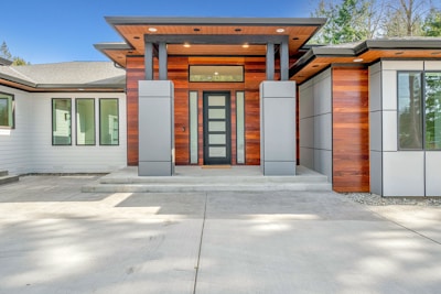 A modern house entrance featuring a balance of wooden and metallic elements. The facade includes large wooden panels alongside gray metal sections, with a central door made of vertical opaque glass panels. The entryway is supported by columns and leads onto a smooth, gray concrete path. Large windows flank the sides, allowing for considerable natural light, and a sloped roof adds to the architectural design.